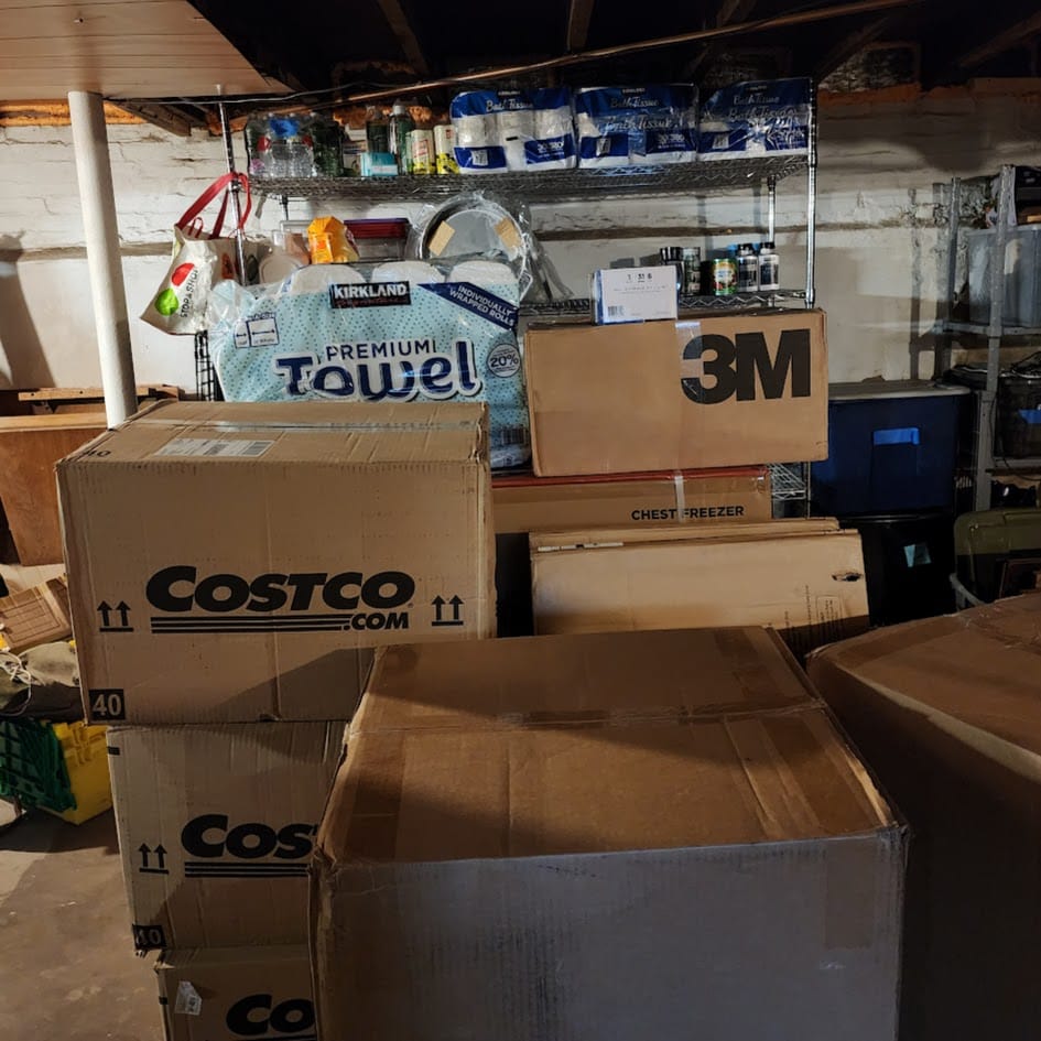 Several cardboard boxes stacked on top of each other in a basement, in front of a storage rack for dry goods.