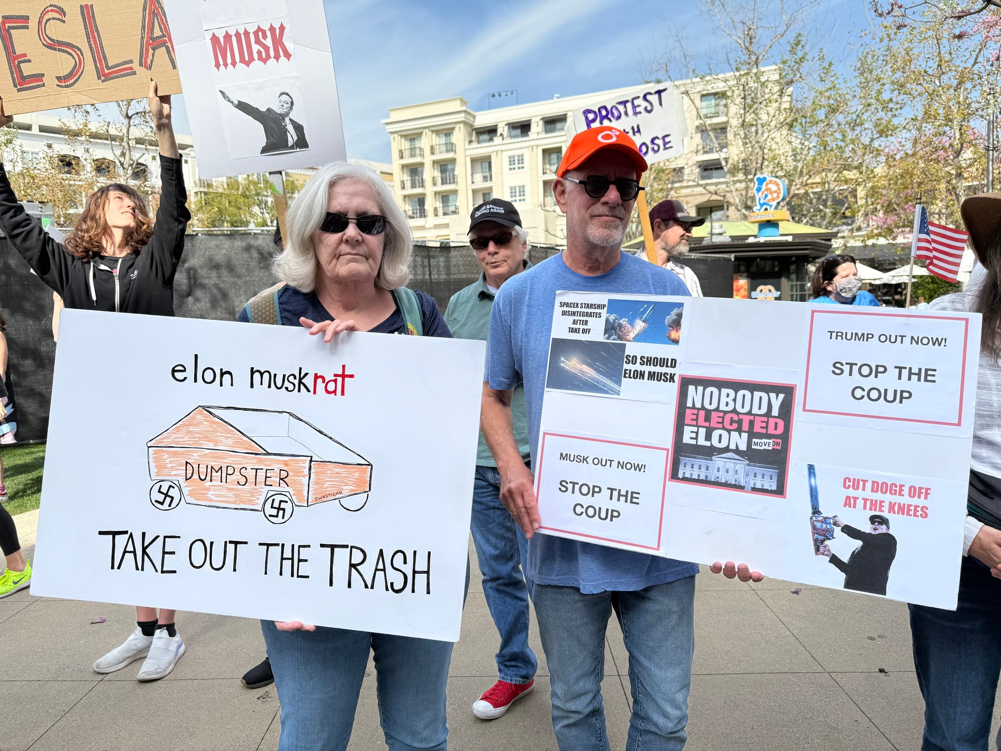 Two people hold signs at the anti-Tesla rally, one says Elon Muskrat, take out the trash with a Cybertruck labeled as a dumpster