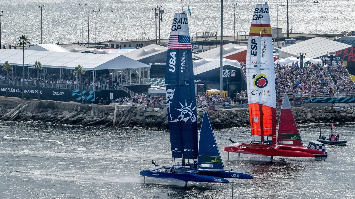 Two boats with USA and Spanish sails head out of LA's harbor past a huge grandstand with fans watching the race