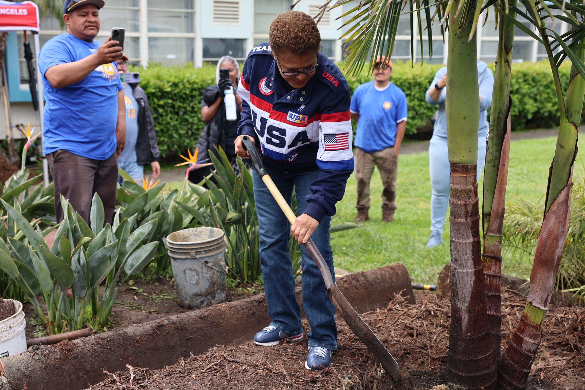 Mayor Karen Bass wearing a Team USA coat while planting a palm tree at a volunteer day