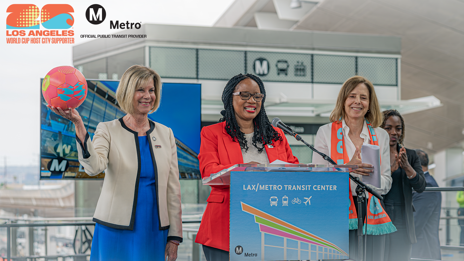 Officials at the LAX Metro Transit Center opening hold a FIFA soccer ball and wear FIFA scarf as they make an announcement at a podium