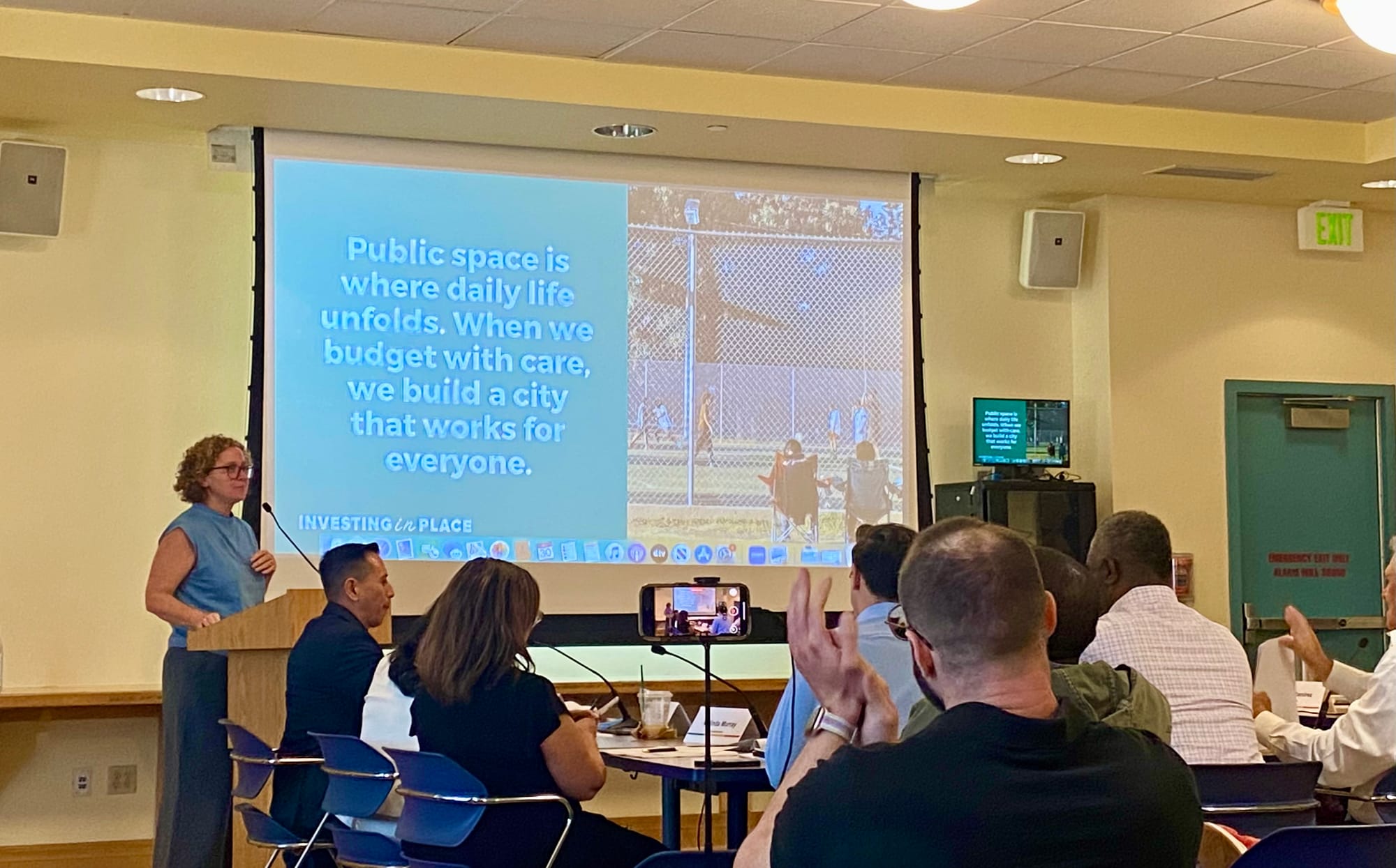 Jessica Meaney standing at a podium in a meeting room with a large screen behind her that reads: "Public space is where daily life unfolds. When we budget with care, we build a city that works for everyone."
