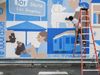 A person on a red ladder painting a mural with a 101 South freeway sign, two people at a cafe, and a streetcar
