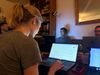 A woman leans over her laptop in a living room of an apartment filled with other people on laptops