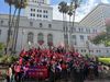 Union members wearing red and purple shirts and holding a sign that says Olympic wage now in front of LA City Hall