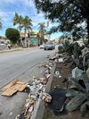 A street filled with trash in the gutter: wrappers, trash bags, cups, buckets, boxes, clothing, car parts
