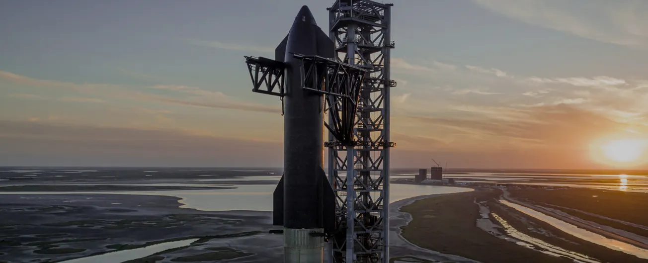 A large steel rocket hangs from the arms of a tall metal tower over the coast of Texas.