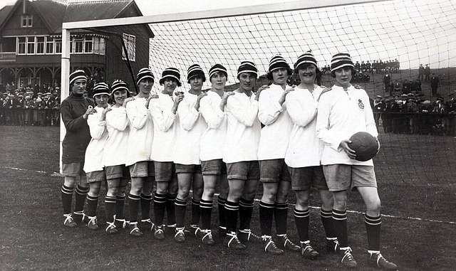 Black and white image of 12 1920s women lined up in front of a soccer net. 11 are wearing white, with striped hats. One is in a dark color..