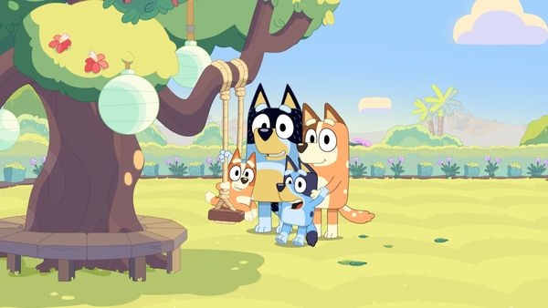 An animation still of the popular cartoon dog, Bluey, with her parents and sister by a tree.