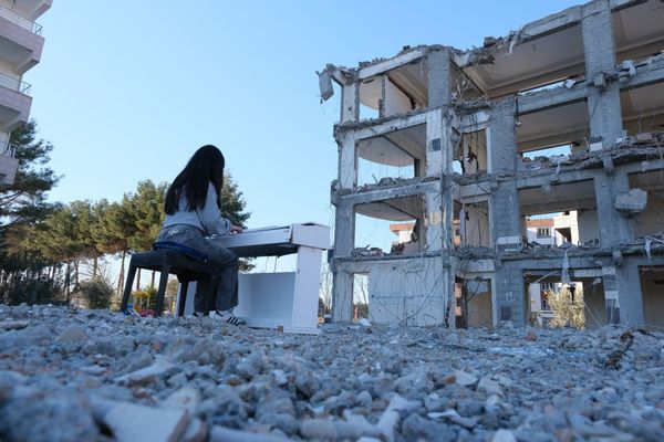 We see the back of Dağdeviren at a piano on a pile of rubble, the shell of a building in front of her. 