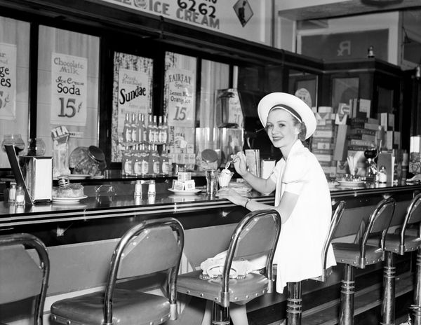 A black and white photo shows a woman sitting at a diner counter eating a sundae by herself. Circa 1930.