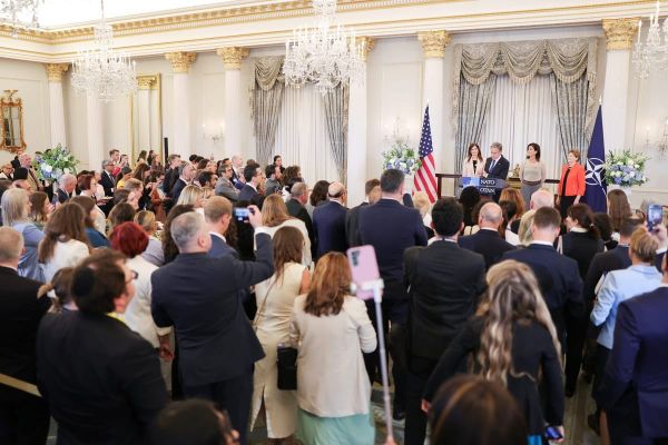 Read view of a packed White House room as Antony Blinken stands at a podium, flanked by women 