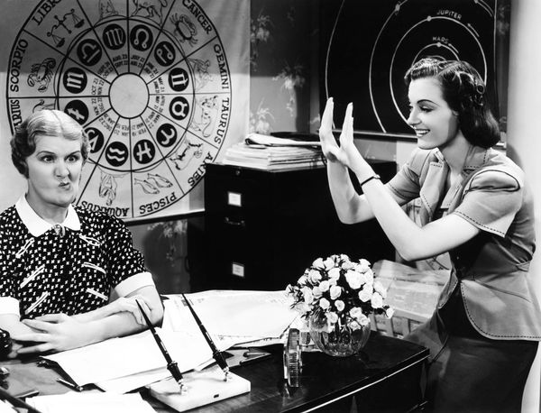 A still from the film "Personal Secretary"—a woman at a desk with a zodiac chart behind her makes a funny face.