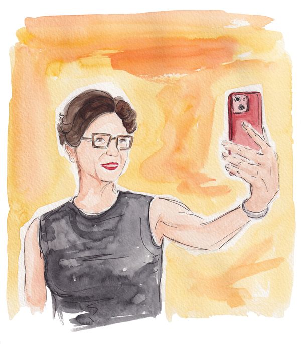 Leora Tanenbaum, a white woman with short dark hair, square glasses, red lips and a black top, smiles into a phone camera.