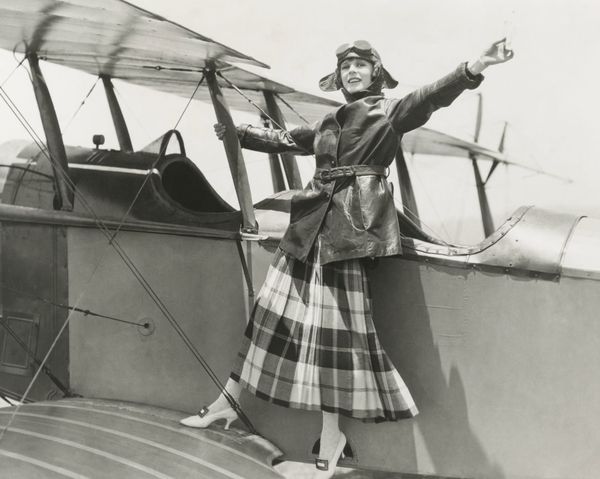 A vintage photo of a woman standing on the wing of a small airplane.