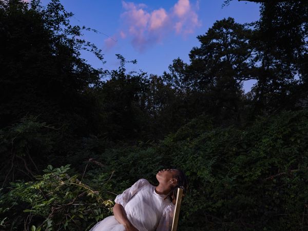 A woman in a white dress, sitting on a chair, twists her body to look up at the sky.