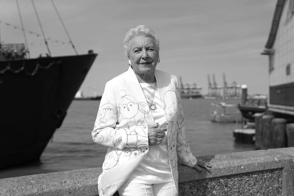 Dressed in a white blazer, white shirt and white pants, Stephanie Shirley poses with boats behind her.