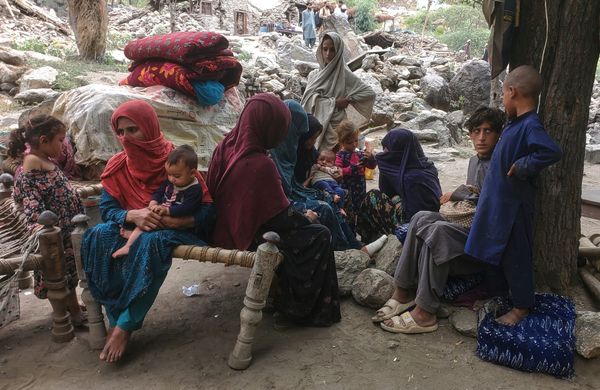 A women with a covered face and a baby on her knee is surrounded by rubble and other survivors. 