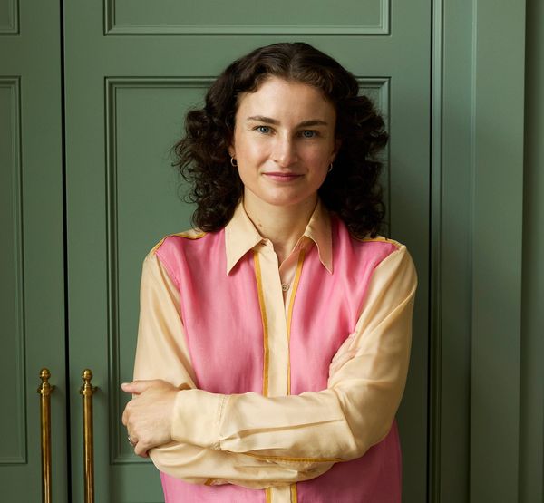 The new editor of Vogue stands against a backdrop of green doors, her arms crossed in front of her. She wears a yellow and pink button-down and jeans.