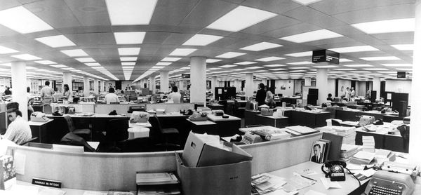 A black and white photo of an old-school newsroom: open seating at long desks, rotary phones, typewriters.