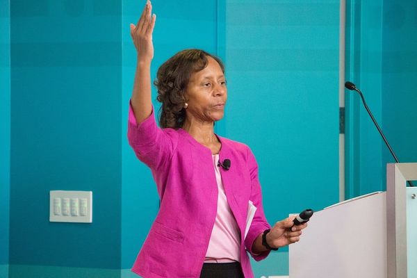 Marian Croak, wearing a pink jacket, waves one hand in the air and holds a microphone in the other. 