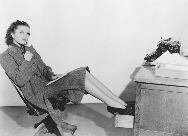 A retro image of a woman in heels sitting at a desk chair, with her legs propped up on a desk drawer.
