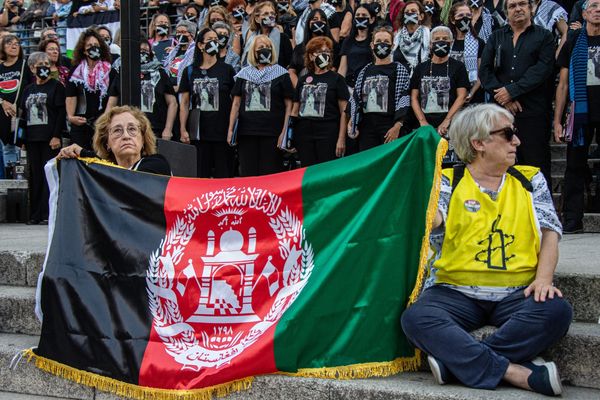 Two women sit cross-legged on steps, holding an Afghan flag. Behind them, women wear face masks to symbolize their silencing