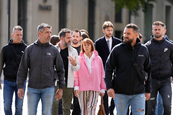 Giselle Pelicot, a small woman in a pink jacket and pastel striped pants, walks through Nimes. She is surrounded by men, all wearing jeans and black jackets.