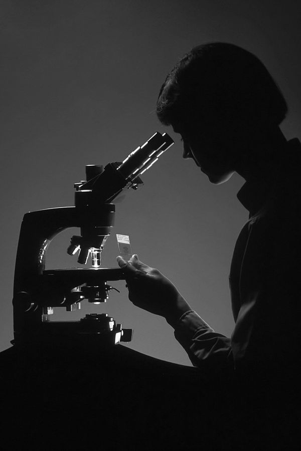 A woman is silhouetted against a blue background as she peers into a microscope. 