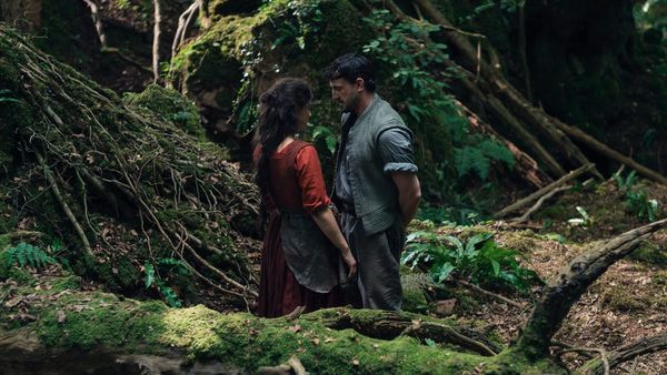In still taken from the movie Hamnet, Agnes and Will stand in a forest, close together, as though they are about to kiss.