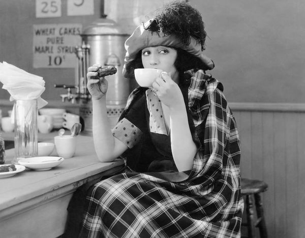 Black and white photgraph of a woman in 1920s-style clothing enjoying a cup of coffee and a cookie at a bar.