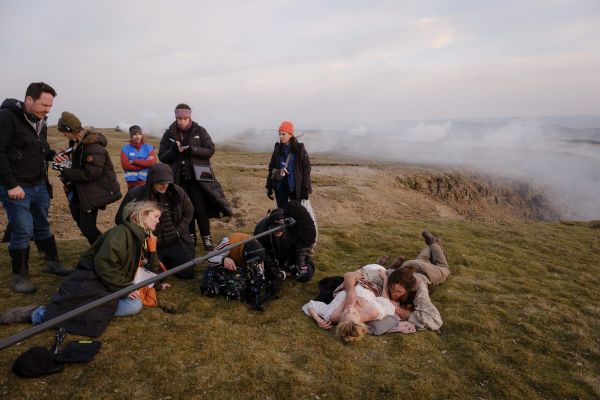 Behind-the-scenes shot of Margot Robbie and Jacob Elordi rolling around on a moor as crew members look on.
