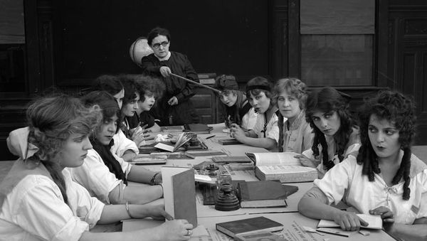 Black and white still from a silent movie showing girls sitting around a large table, reading books.