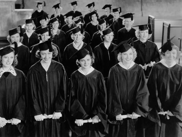 Black and white still of several rows of white women, who look to be from the 1950s, in caps and gowns holding diplomas.