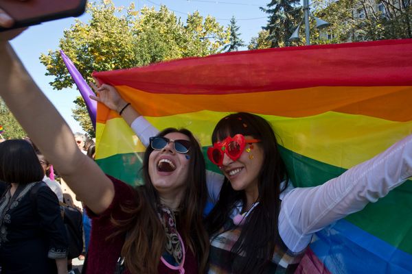 Two people with sunglasses and long hair standing in front of a Pride flag. They are smiling.
