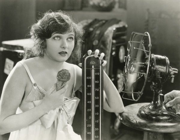 A still from an old movie shows a woman with a fan and melty ice cream cone, hanging on to a thermometer.