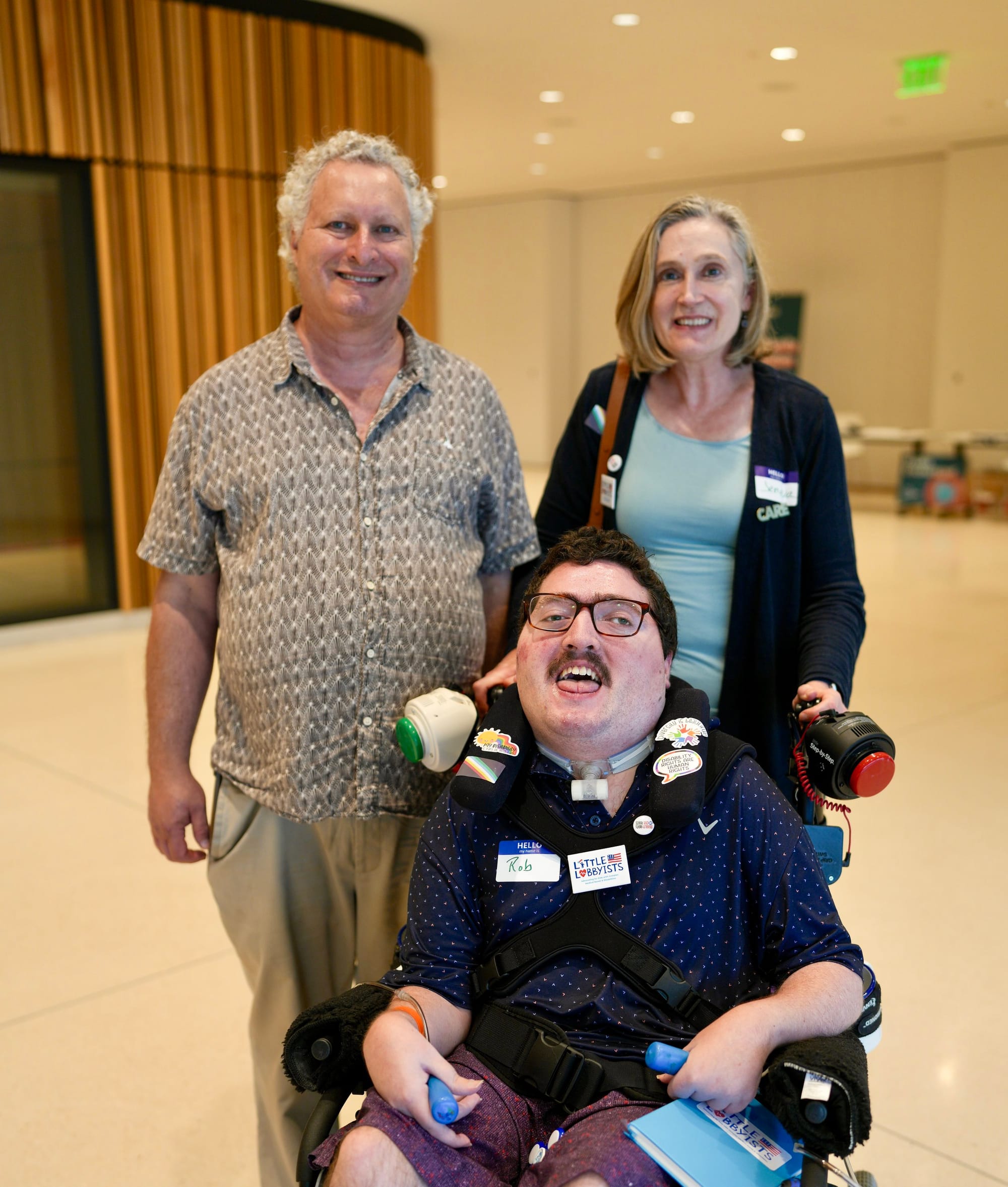 Little Lobbyists Roger, Jeneva and Rob pose inside the conference area in the MLK library. Rob is using a wheelchair and his vest is covered with disability advocacy pins. He is holding his AAC communication switches. Photo credit: Rah Studios 