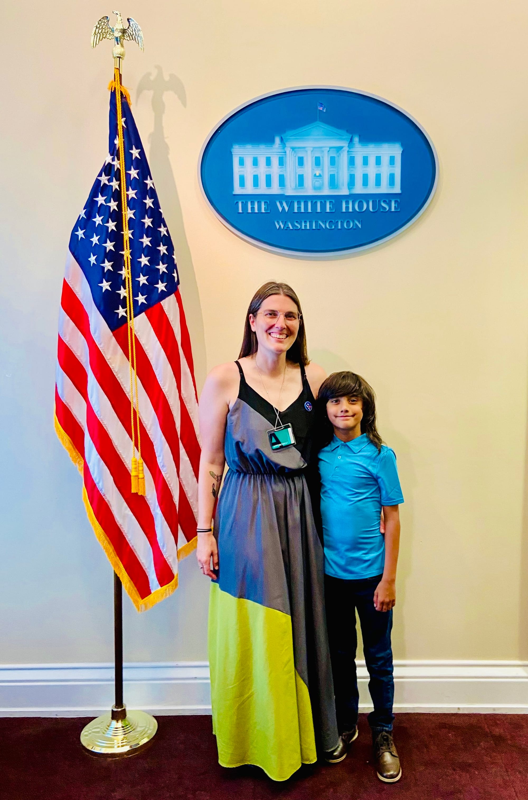  Little Lobbyists Ali and Ethan pose in the Old Executive Office building in front of a wall with the White House logo (blue &amp; white oval) above them and a U.S. flag to the left. Ali wears a long skirt and Ethan is dressed more casually. 