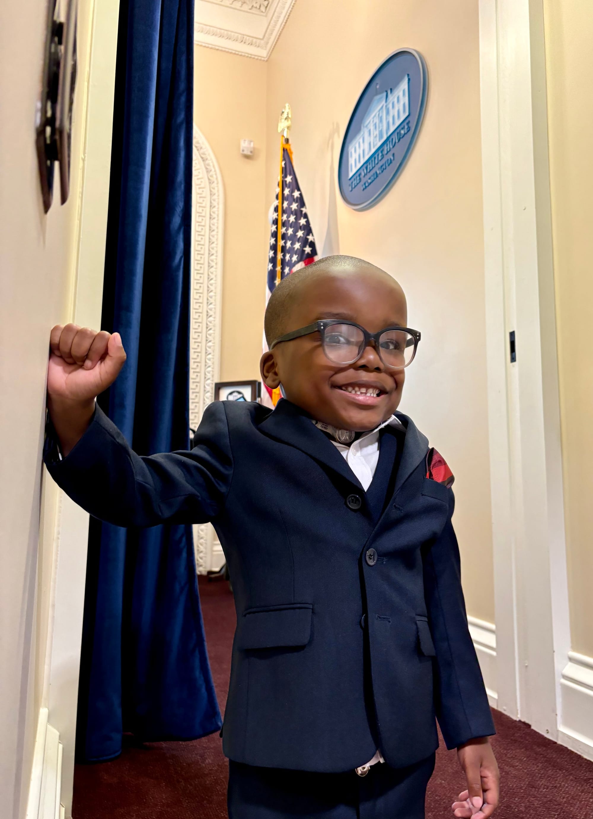  Adonise wearing a sharp, tailored suit coat, poses in a hallway in the OEB, with a White House logo to his right on a wall. 