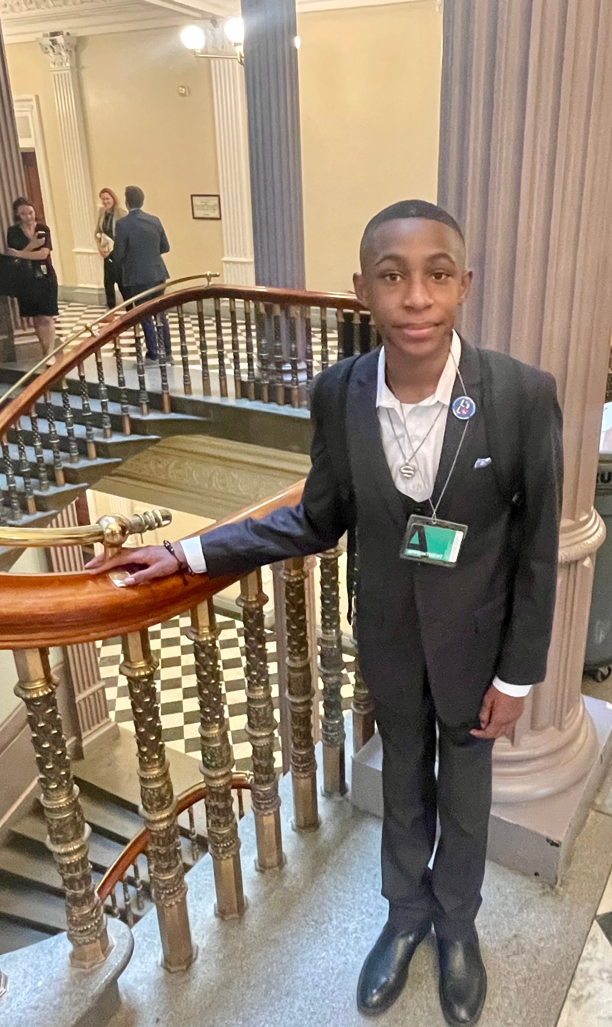  Isra'El wears a tailored suit coat and pants, with a white button-down shirt. He stands in a stairwell in the OEB, his hand on a decorative metal railing. The 19th century columns and other architectural features of the historic building are behind 
