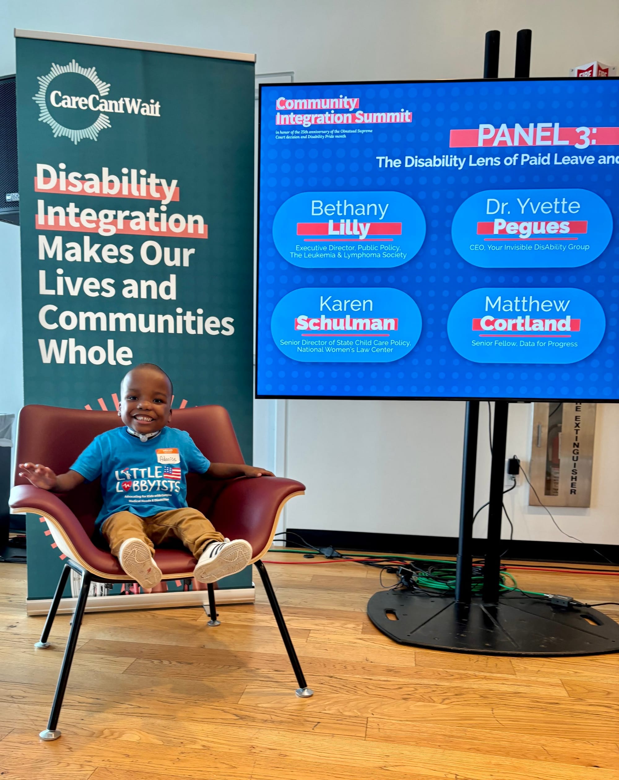  Adonise sits in a chair in the conference area of the library. Behind him is a banner with the Care Can't Wait logo and the summit title, "Disability Integration Makes Our Lives and Communities Whole." An electronic board is to the right, giving the