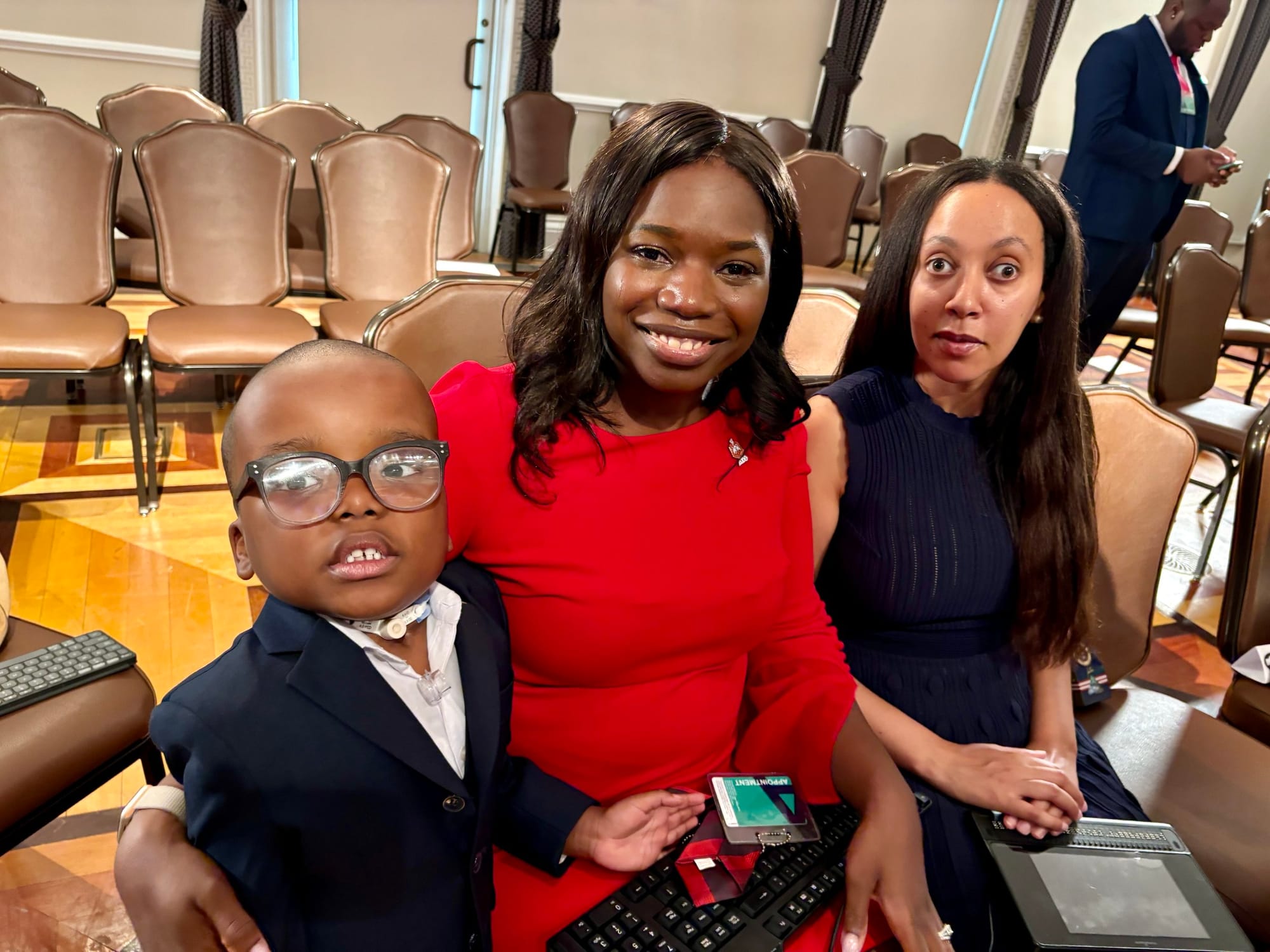  Adonise and his mom Anne are seated in a presentation room in the OEB. Disability advocate Haban Girwin is seated next to them. Anne wears a vivid red dress and Adonise wears a suit. 