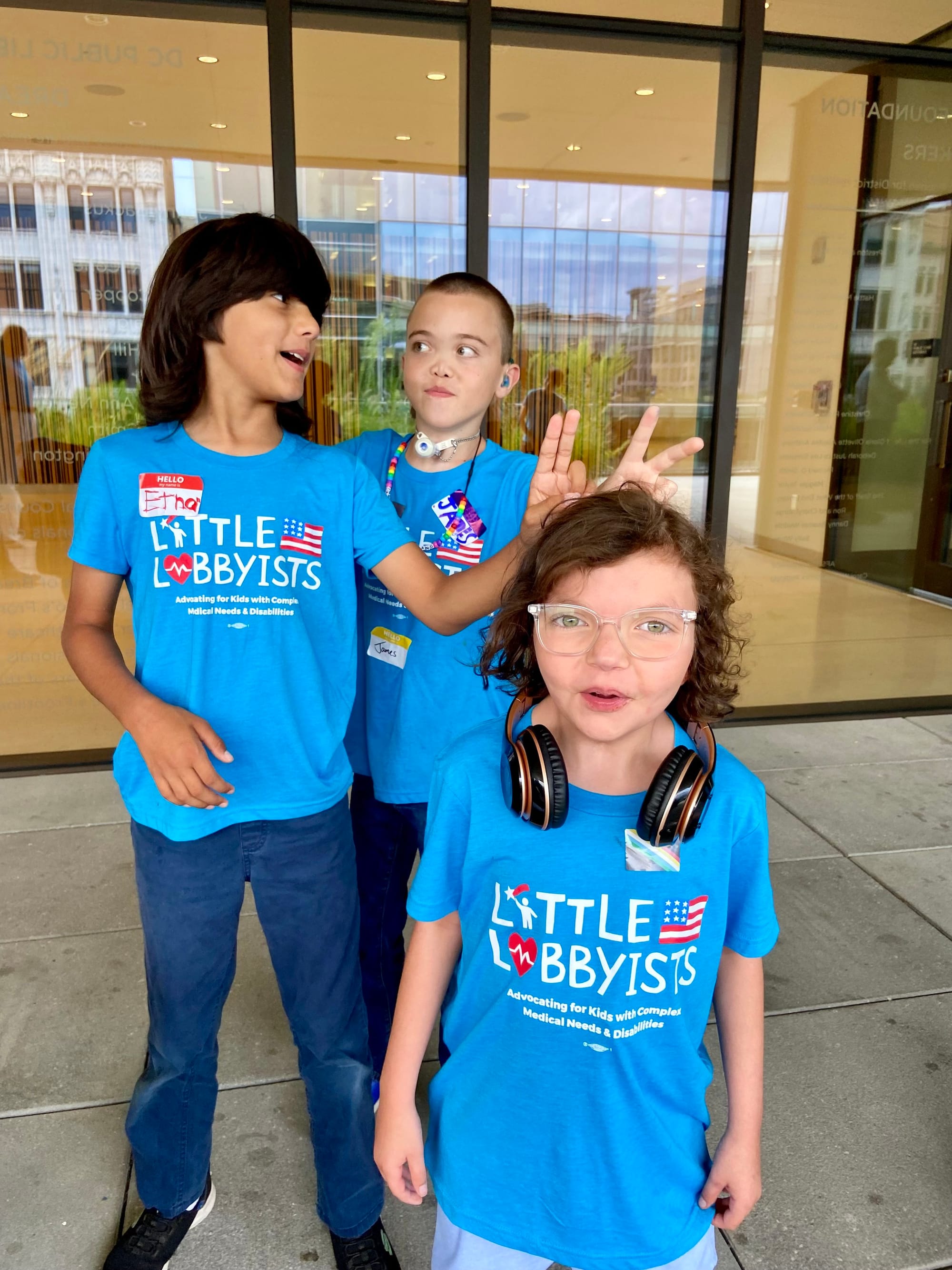  Ethan, James and Louisa, wearing turquoise LL t-shirts, pose on the library's rooftop plaza, under an overhang, with plateglass windows behind them. 