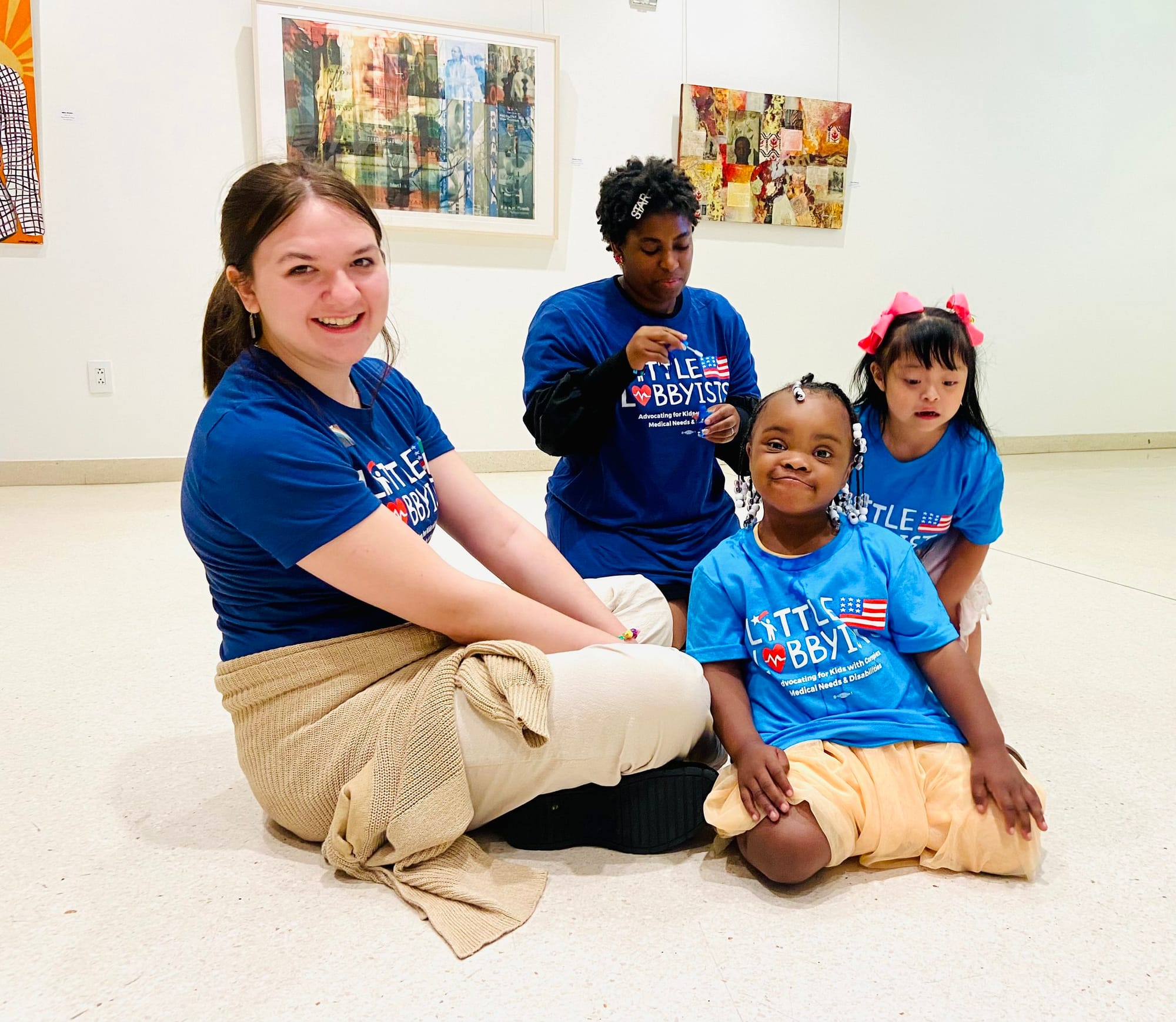  Olivia poses with Destiny and others inside the conference area of the library. All are seated on the tiled floor and framed artwork hangs behind them on the walls. 
