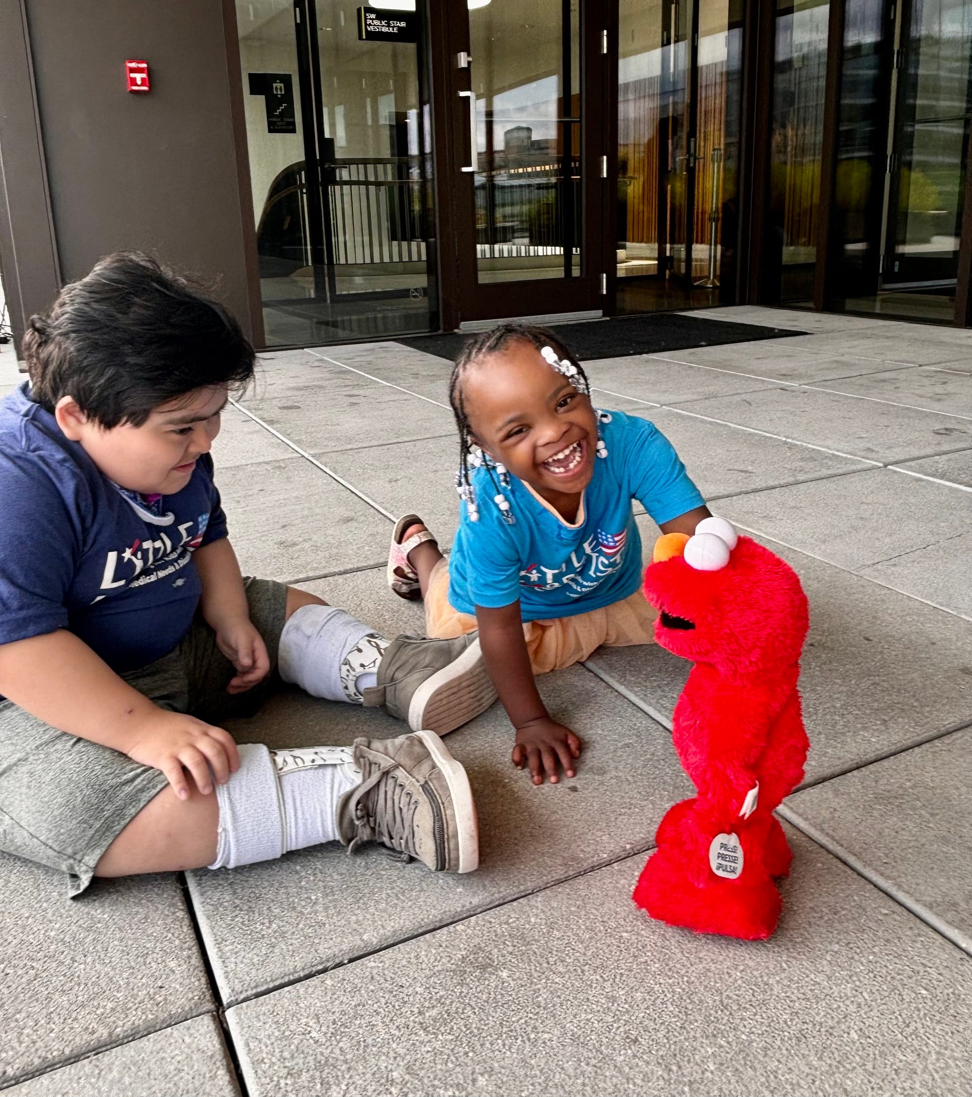  Teddy and Destiny play with a bright red Elmo doll on the concrete flagstones of the library's outdoor plaza. They are smiling and laughing. 