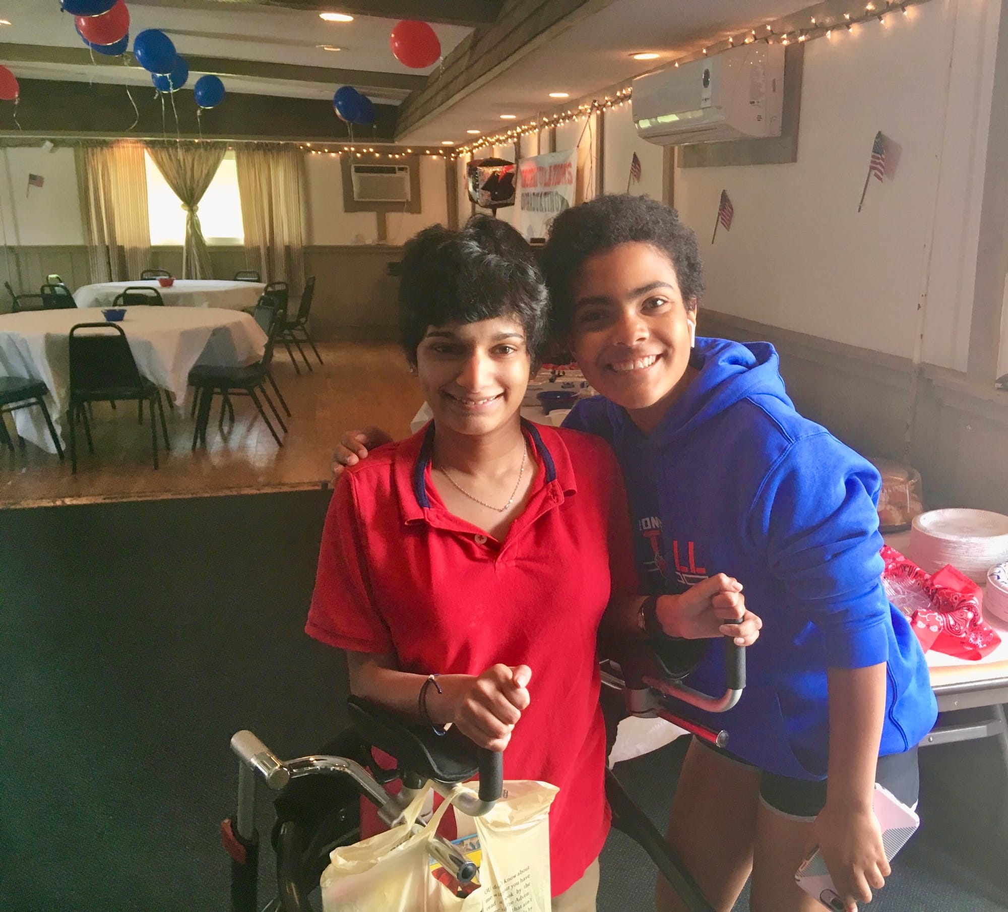Grace Dow (r) with her young sister (l) at Grace’s high school graduation party. Grace wears a red shirt and her sister a blue shirt. Both women have dark skin and dark hair. They are in a room set up for a party with red and blue balloons and tables