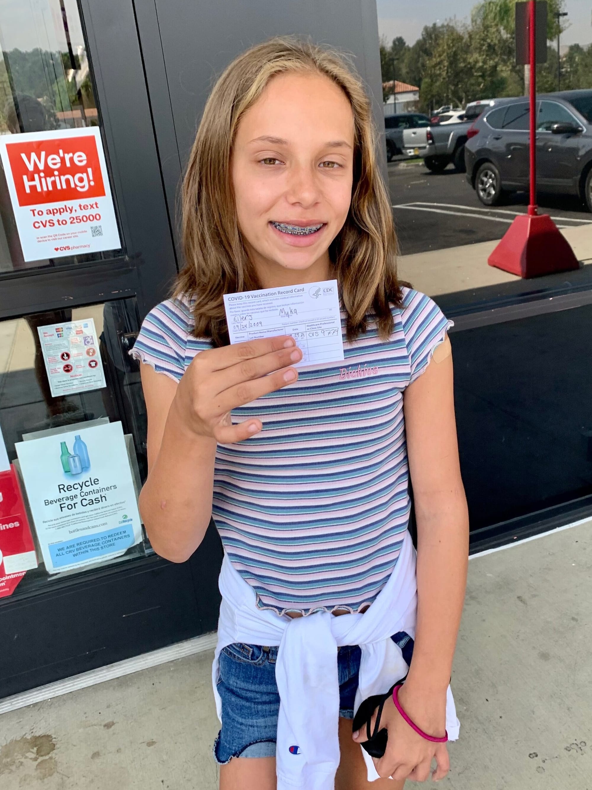 Myka holding her vaccine card. [image description: A light-skinned girl with shoulder-length blond hair holds up her vaccination card. She is standing outside a chain drug store, with signs on its windows. She wears a striped t-shirt and denim shorts.]
