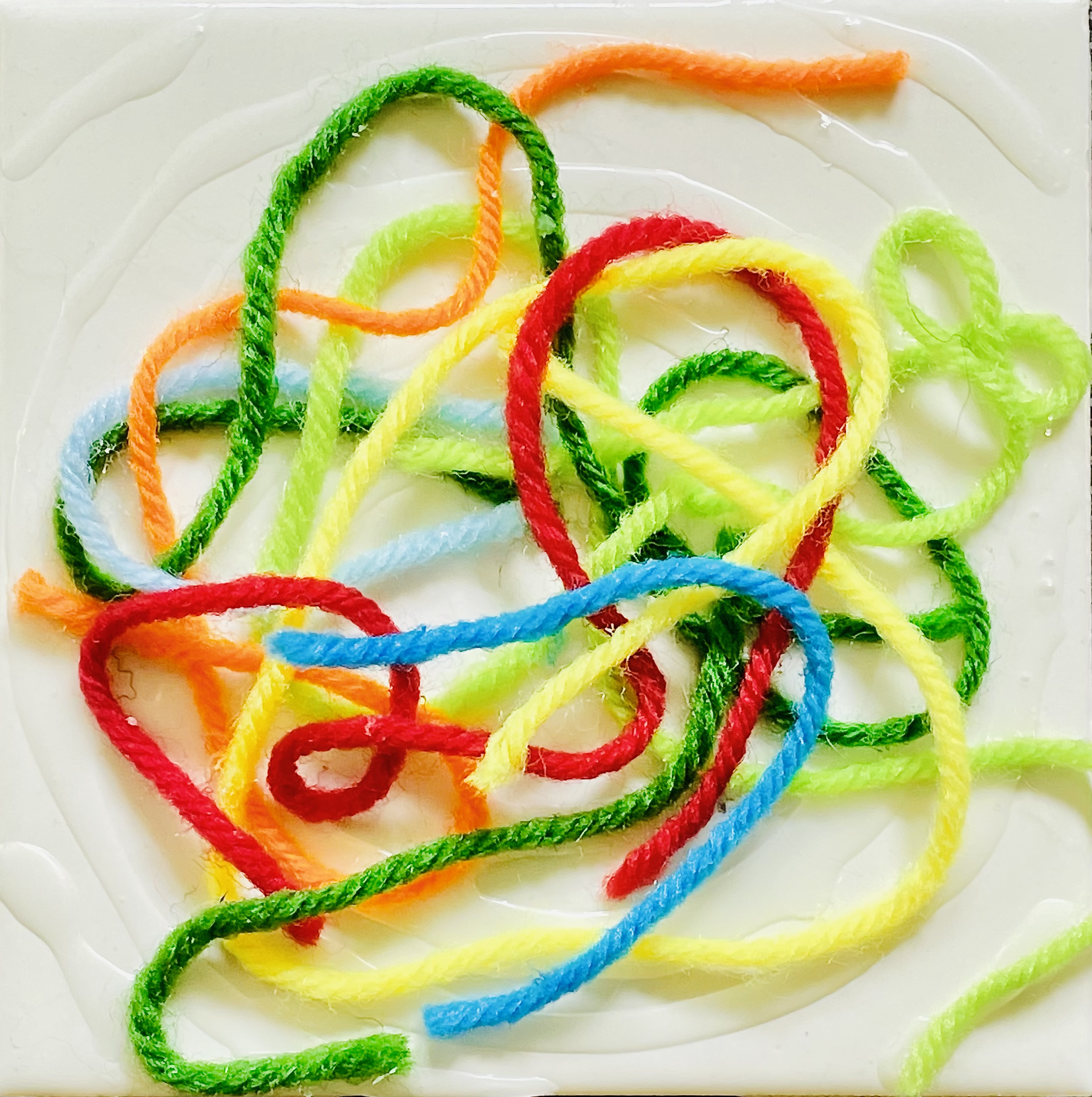 Art by Rob Stone, yarn on ceramic tile. [image description: Pieces of yarn (dark &amp; light blue, light &amp; dark green, red, orange, and yellow) have been dropped in tangled loops on a glossy white ceramic tile and glued into place.]