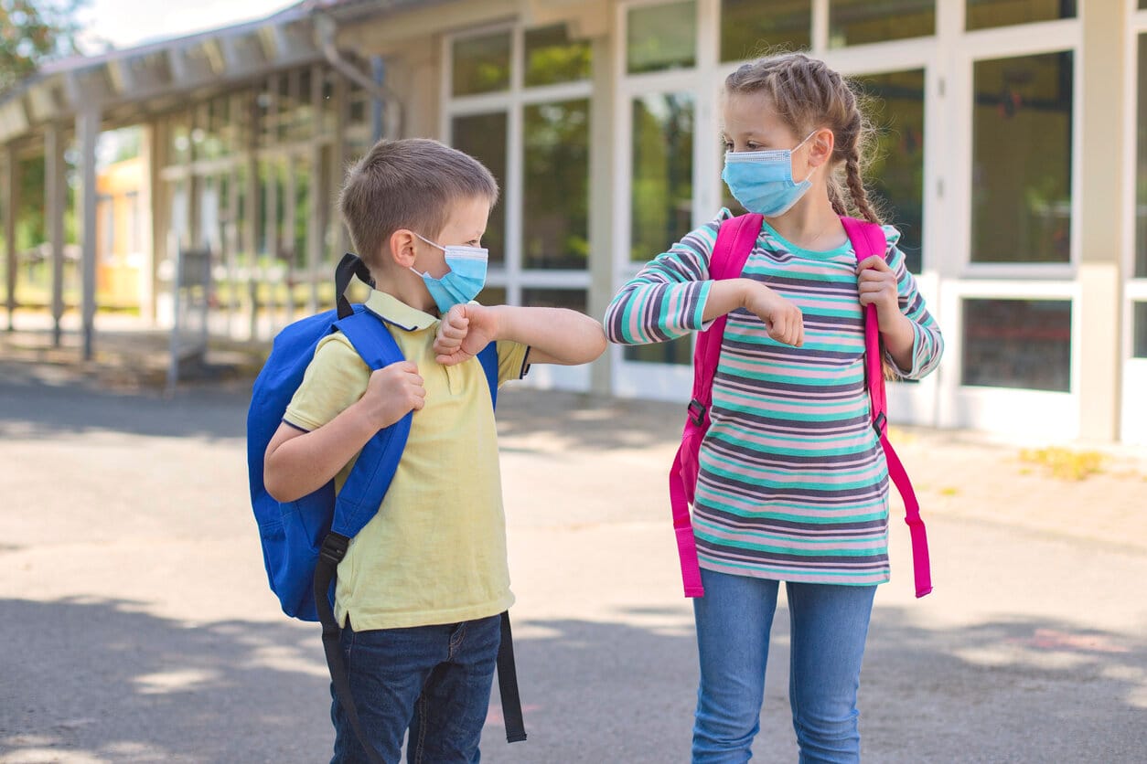 Stock photo. [image description: A young boy (left) with pale skin wears a blue surgical face mask and yellow shirt and carries a royal blue backpack. He elbow-bumps a young girl (right) wearing an identical face mask who has pale skin and braided hair. She wears a blue-striped shirt and carries a pink backpack. The two stand in front of a school building.]