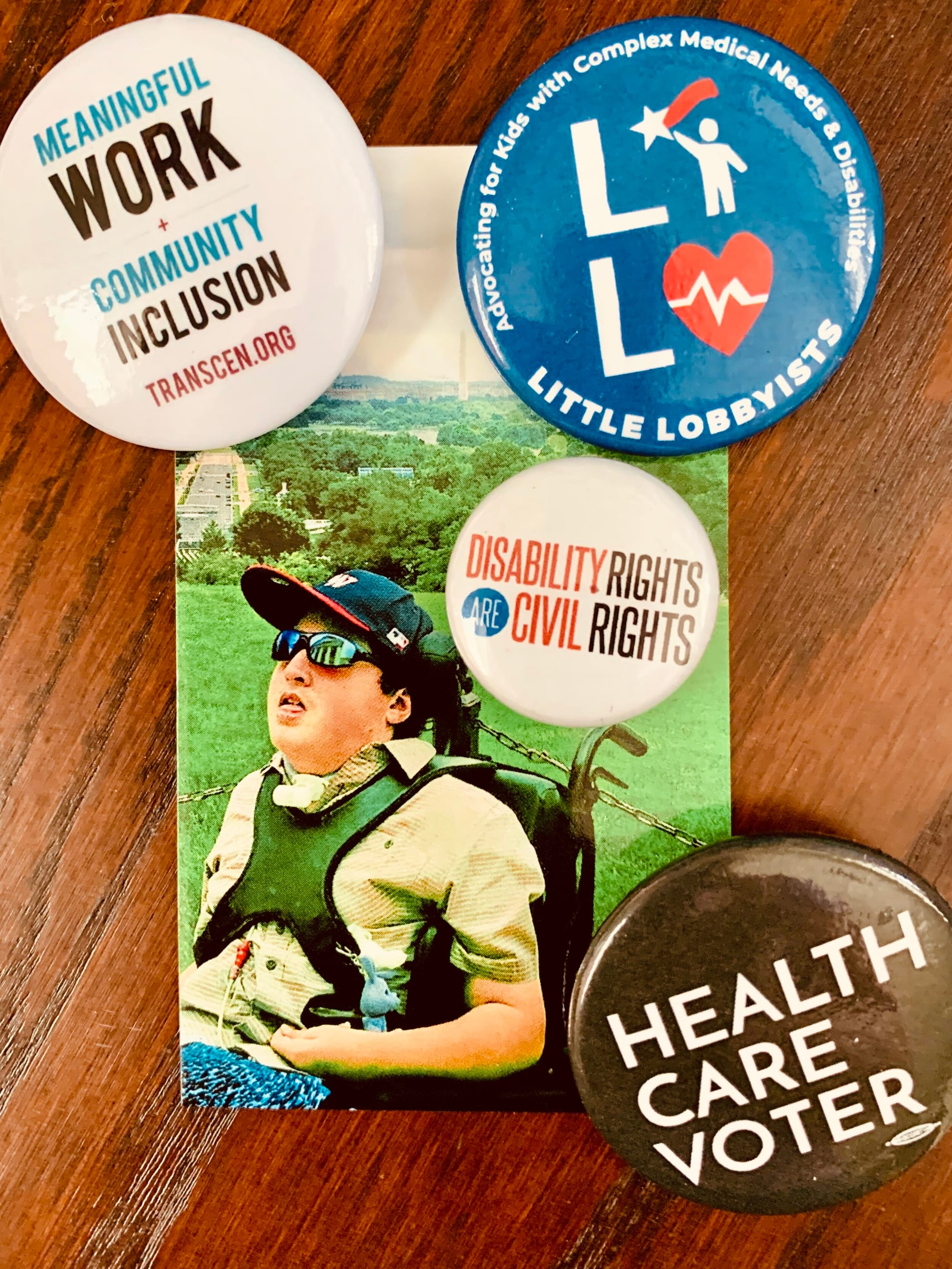 Rob and his disability rights button collection. [image description: A business card on a brown wood table shows a man with light skin sitting in a wheelchair on a grassy hill overlooking the city of Washington DC. He wears sunglasses and a Washington Nationals baseball hat. Four round advocacy pins are placed near the business card: “Meaningful Work Community Inclusion Transcen.org; Little Lobbyists (with their star child &amp; heart logo), “Disability Rights are Civil Rights”; “Health Care Voter.”]